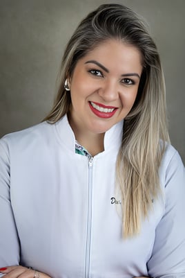 mulher loira com batom vermelho sorrindo, vestindo jaleco médico, posando pra foto em consultório
