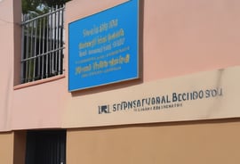 A large auditorium is filled with seated students dressed in uniforms, most of whom are wearing lanyards with identification cards. At the front, there are several individuals, including an older woman in a yellow saree and three men, sitting in blue chairs. The room is well-lit with ceiling lights and has decorated walls.