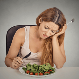 Uma mulher com expressão desanimada olhando para um prato de folhas verdes e tomates.