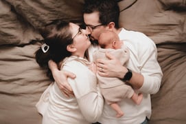 Mom and dad cuddling in bed with their newborn, looking at each other lovingly from above.