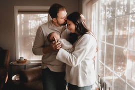 Mom holding and looking at their newborn while dad kisses her head in front of a window during a Gra
