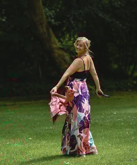  Das Bild zeigt eine lächelnde Frau in einem farbenfrohen, bodenlangen Rock mit floralen Mustern, der Pink-, Lila-, Türkis- u