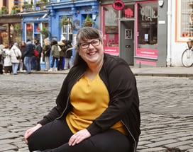 Laura is laughing at the camera sat on a cobbled street with her legs curled up under her
