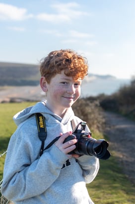 a profile photo of Drew Walker, smiling holding a Nikon D3300 with a background of Cuckmere Haven and the seven sisters.