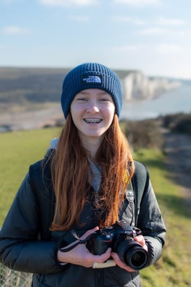 a profile photo of Lilie Walker, smiling holding a Nikon D5300 against a backdrop of Cuckmere Haven and Seven Sisters