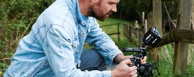 Mann mit Bart in blauer Jeansjacke, draußen, hockend, mit Kamera Rig in den Händen, seitliches Profi
