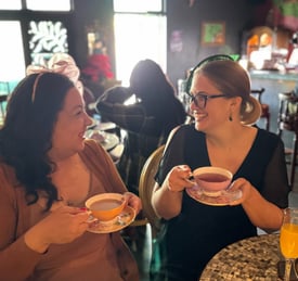 Two women are drinking tea and look at each other smiling. They both are wearing cute little hats.
