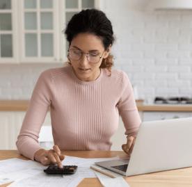 A woman sitting at her kitchen counter working on her calculator and laptop.