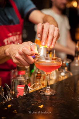 Ein Mixologe erhitzt eine Orangenscheibe mit einem Feuerzeug über einem Cocktail.