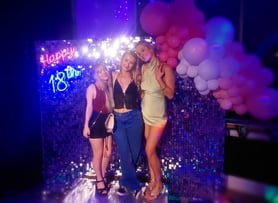 3 girls posing for a photo in front of a silver shimmer backdrop and balloon garland with Happy 18th neon sign hire Brisbane.