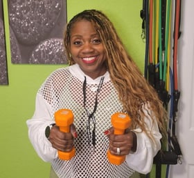 Image of Coach C smiling in workout gear, with two weights in her hands. 