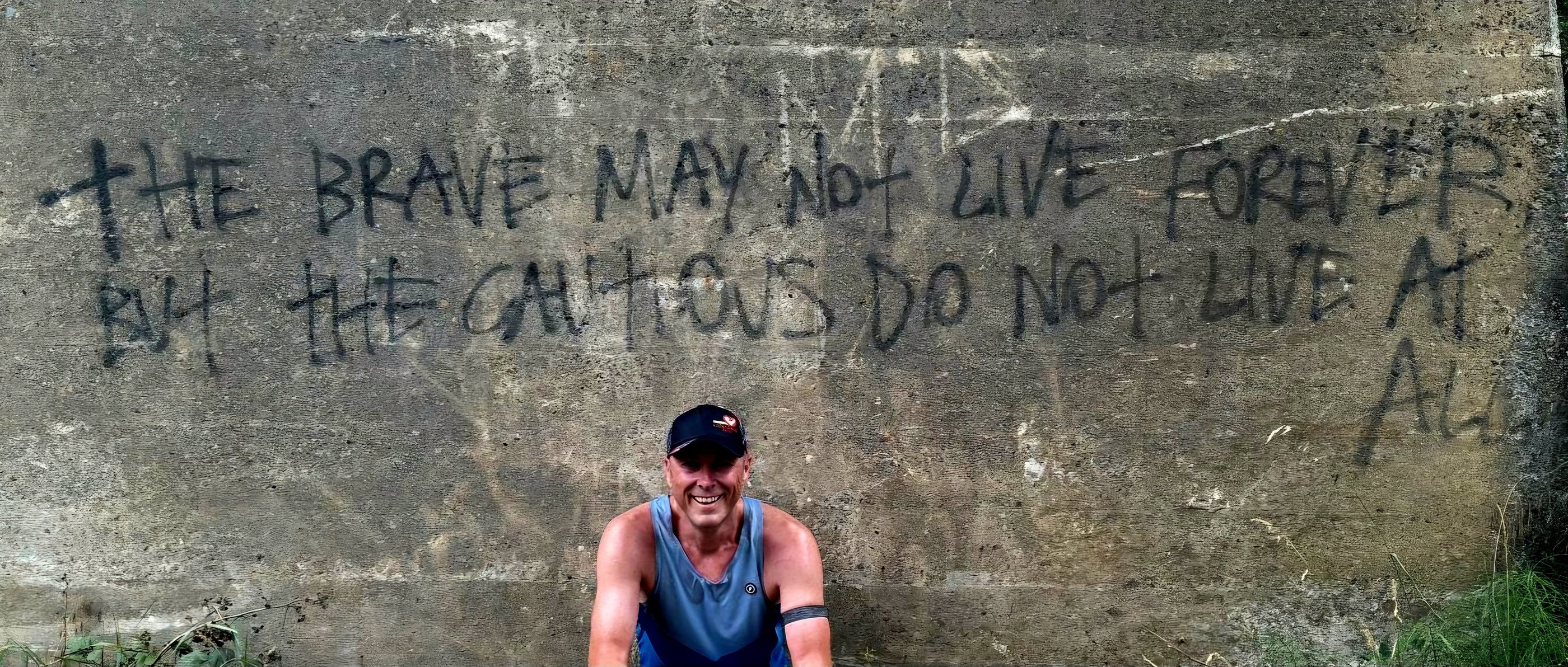 A man smiling in front of an inspirational graffiti quote on a concrete wall about bravery and living life.