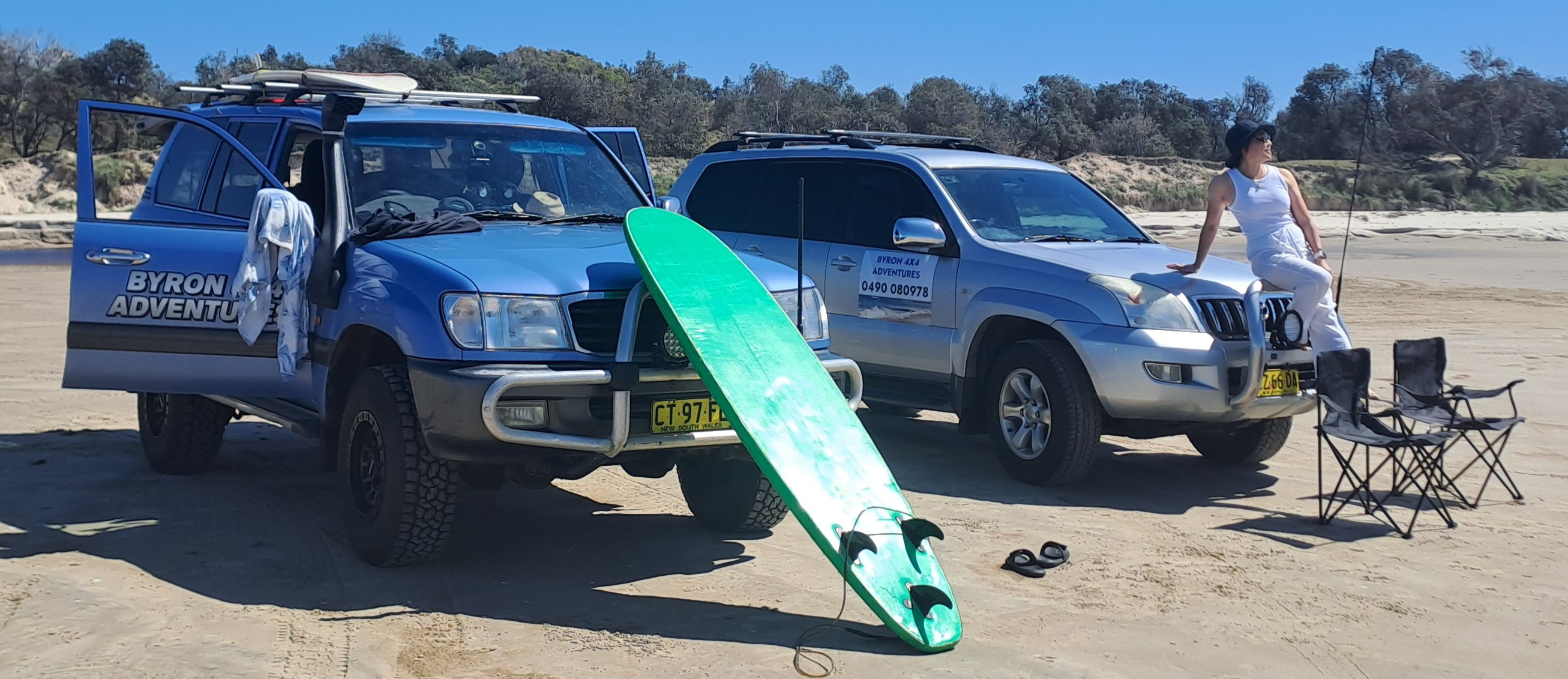 Two 4x4 off-road vehicles parked on a sandy beach with a green surfboard and camping chairs for Byron Bay adventures.