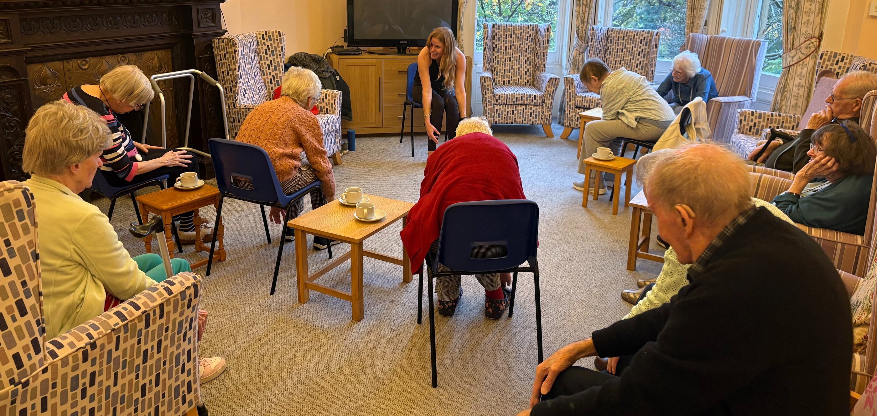 Group of elderly residents taking part in a seated exercise and chair yoga class, led by Lucy