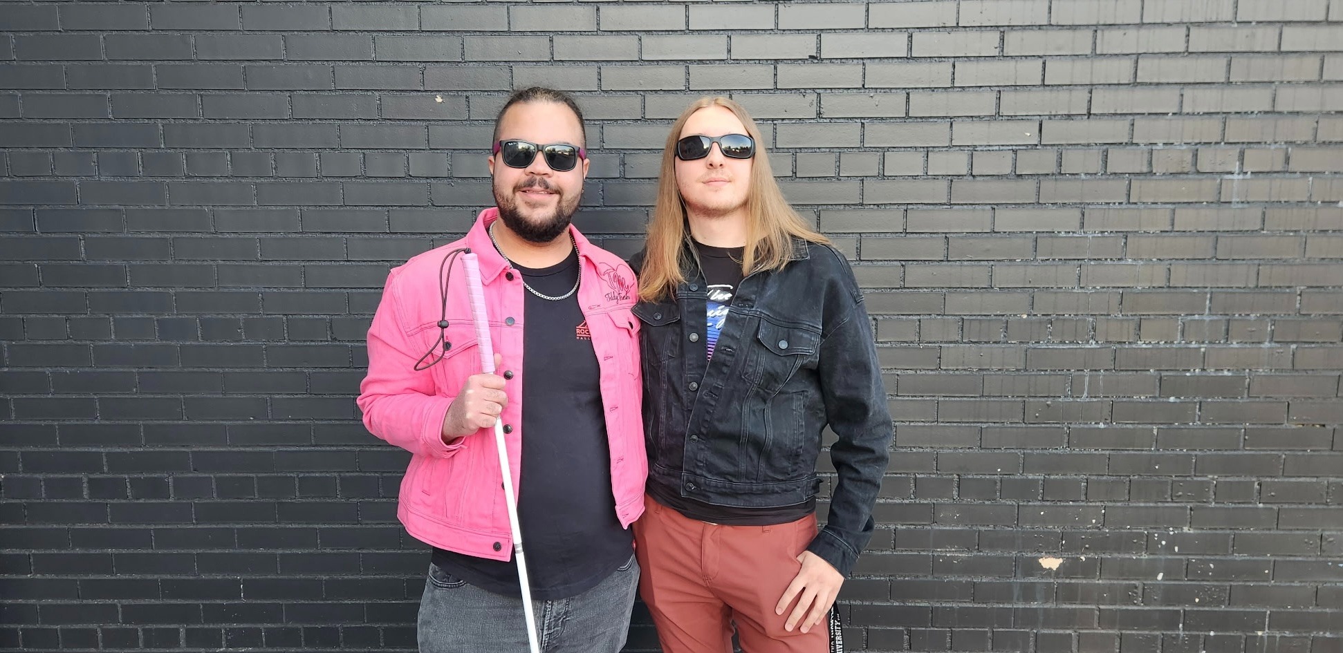 Two men in sunglasses posing against a black brick wall, one holding a white cane for the blind.
