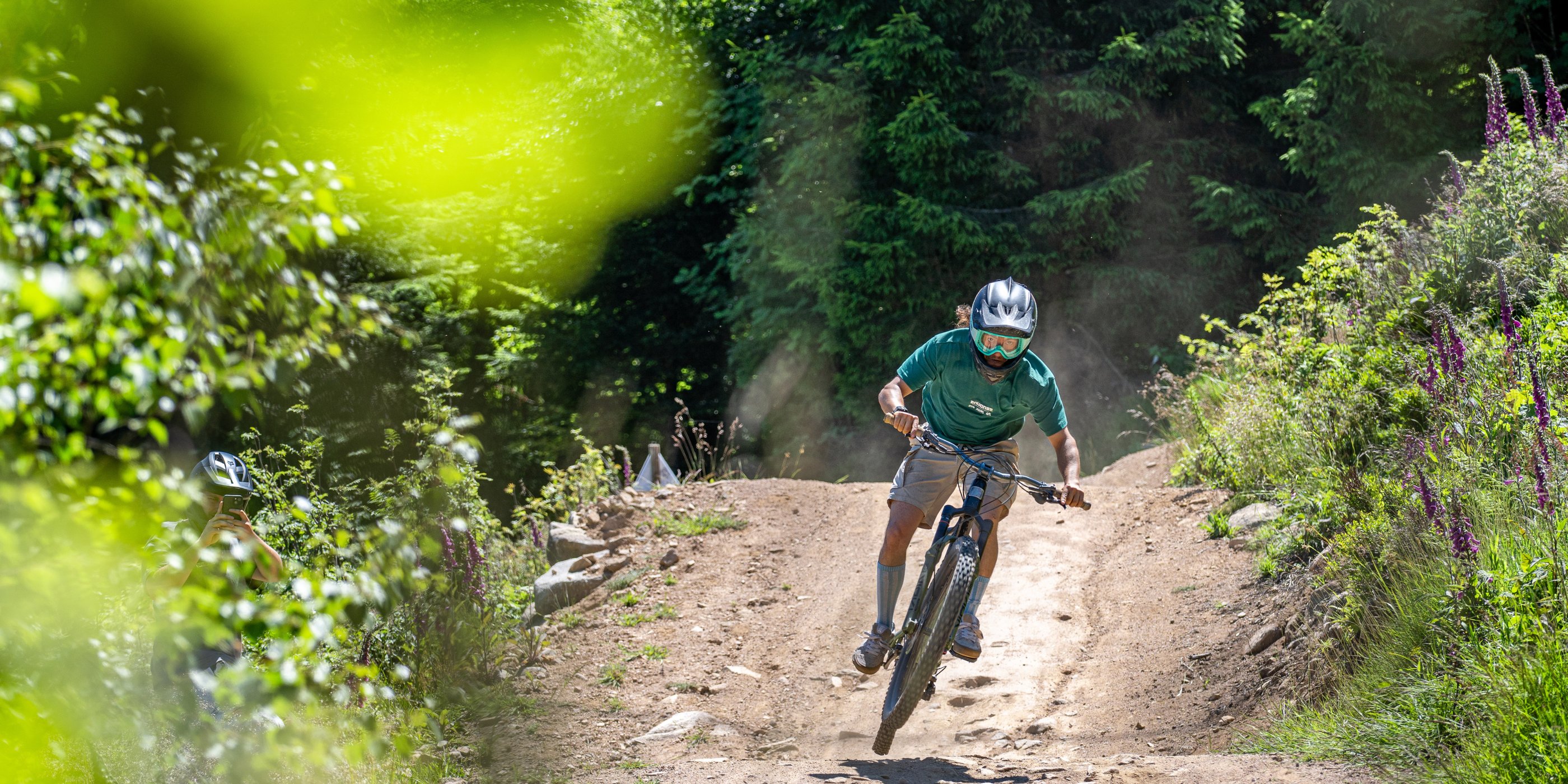 A mountain biker wearing a helmet and goggles jumps over a dirt trail in a green forest.