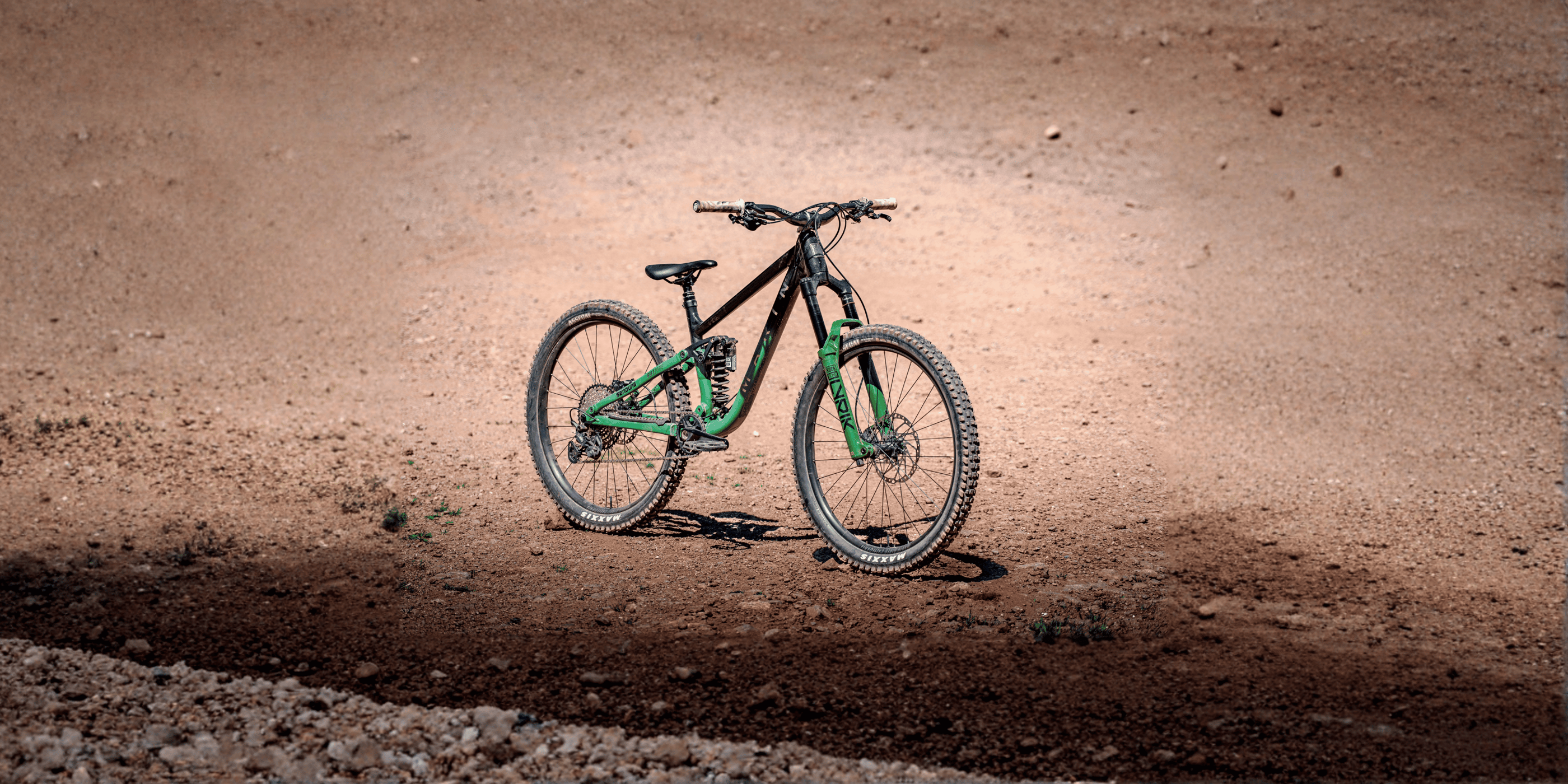 A green and black full-suspension mountain bike parked on a dirt trail at a bike park.