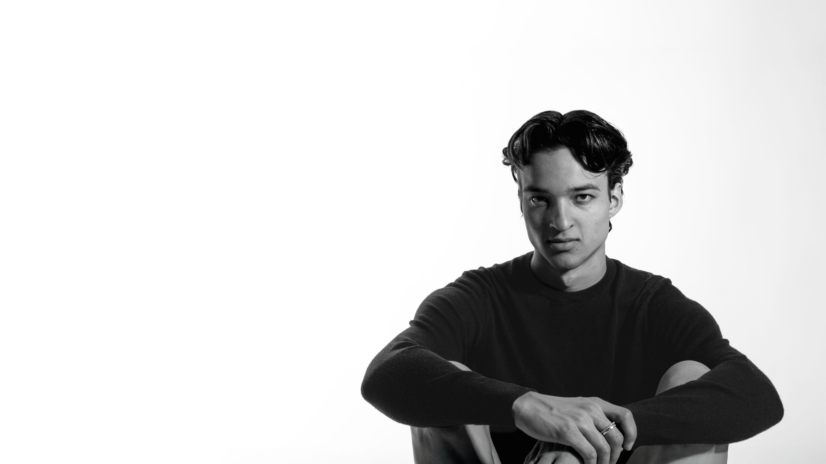 Black and white portrait of a young man in a dark sweater sitting against a white background.