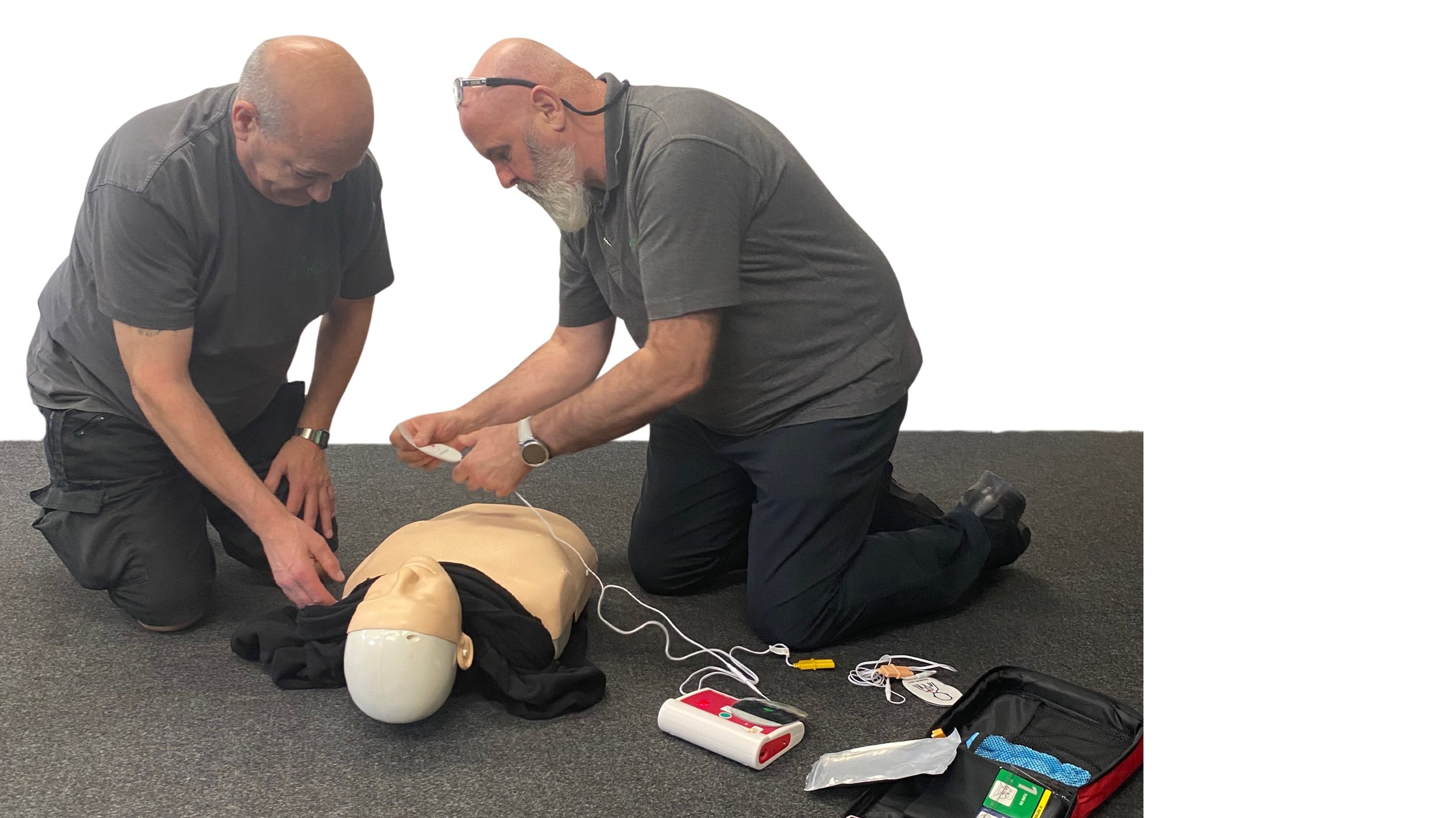 2 men knelt over a training adult manikin, the manikins shirt is raised, AED pads are being attached