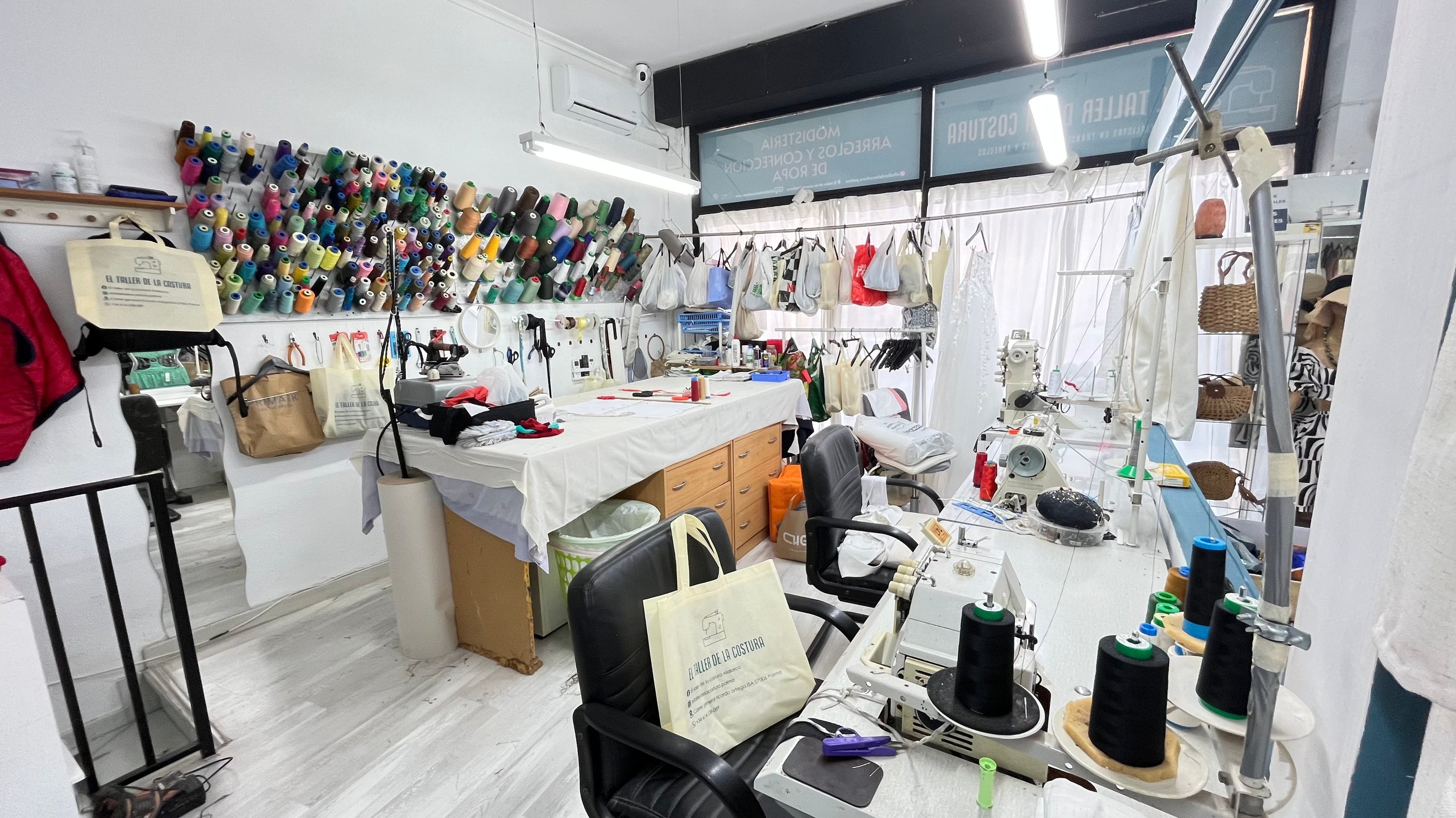 Vista general de un taller de costura con varias máquinas de coser, mesa de corte, rollos de tela, h