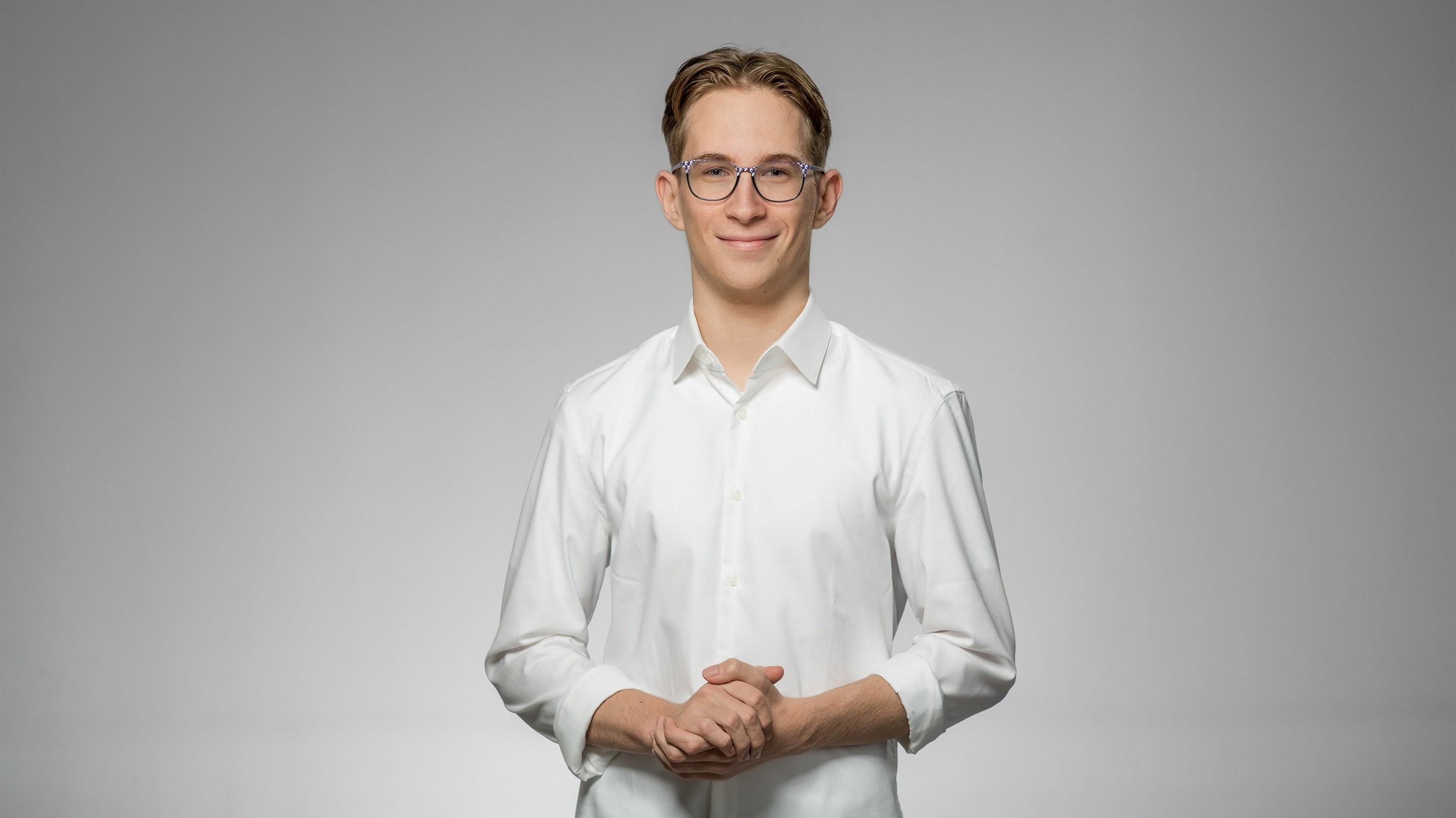 Junger Mann mit Brille in weißem Hemd lächelt freundlich vor neutralem Hintergrund