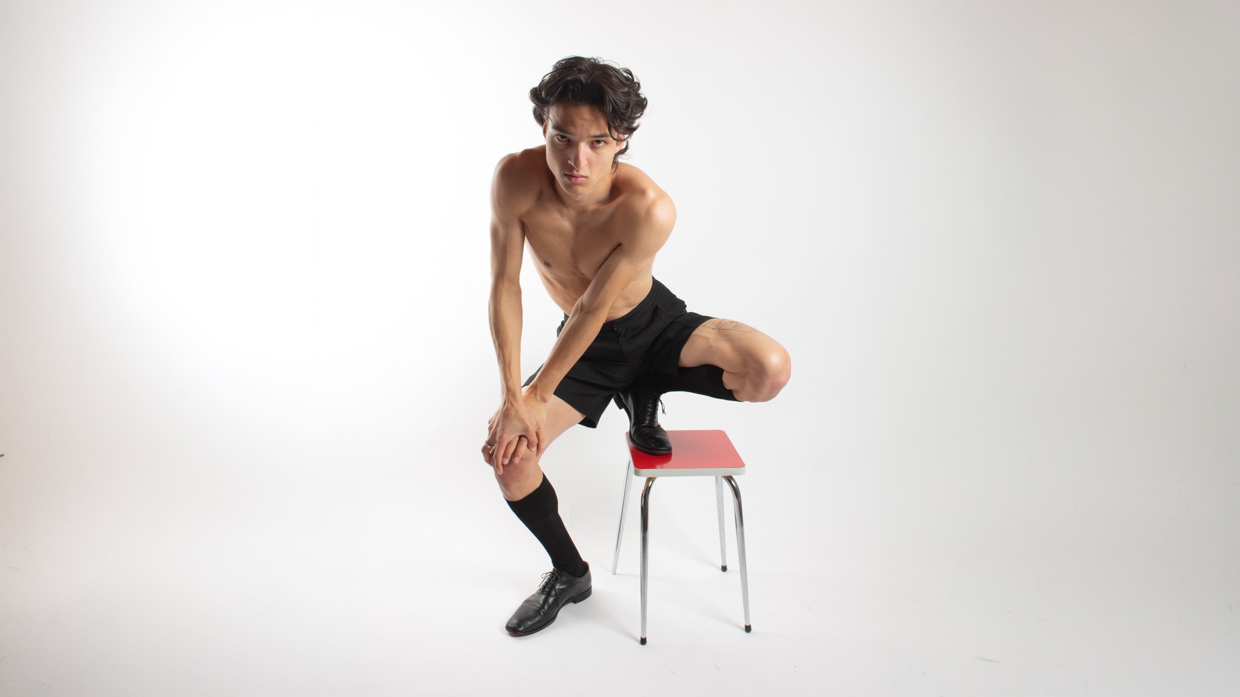 Shirtless male model in black shorts and dress shoes posing on a red stool in a studio.
