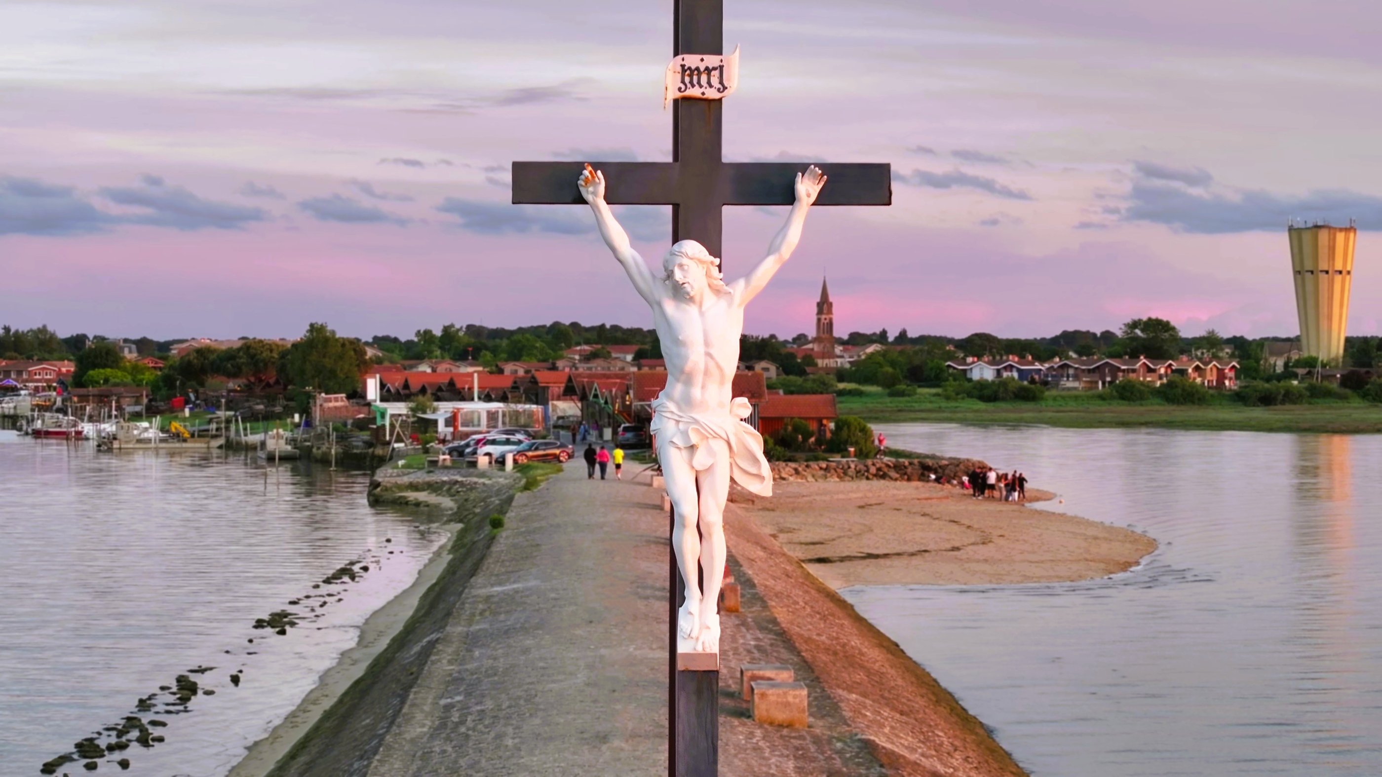 Statue de crucifix blanc surplombant un village côtier et l'eau au coucher du soleil.