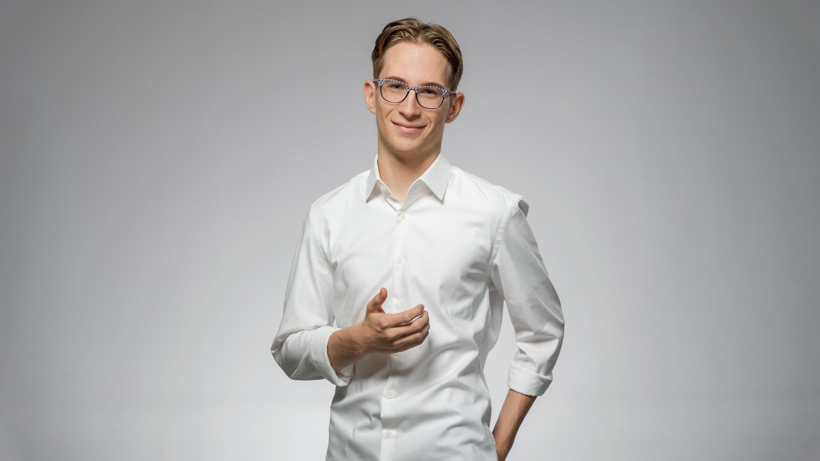 Junger Mann mit Brille in weißem Hemd lächelt freundlich vor neutralem Hintergrund