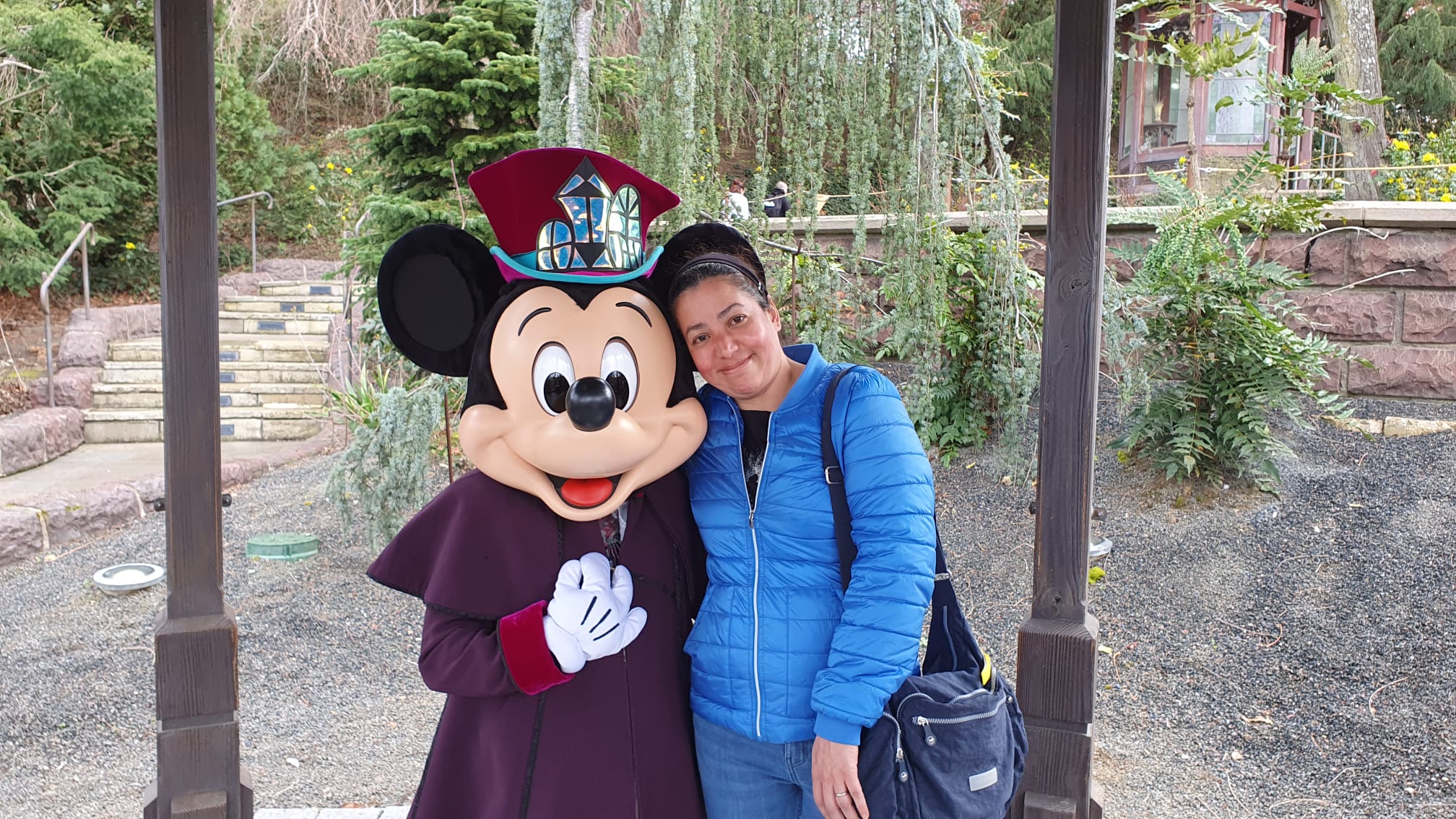A smiling woman in a blue jacket poses with Mickey Mouse in a purple costume at a Disney park.