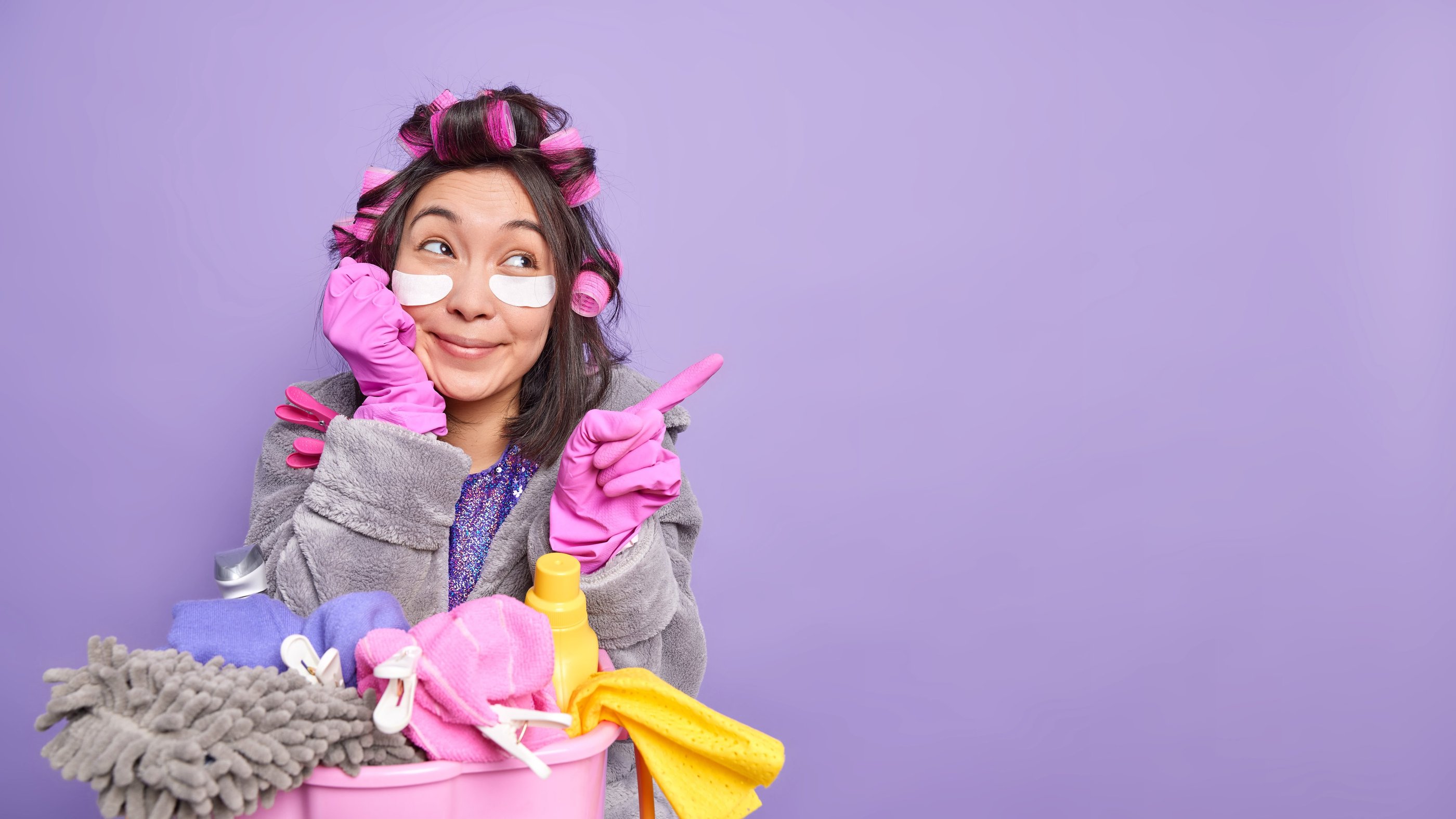 A woman in hair rollers and eye patches wearing rubber gloves with cleaning supplies against a purple background.
