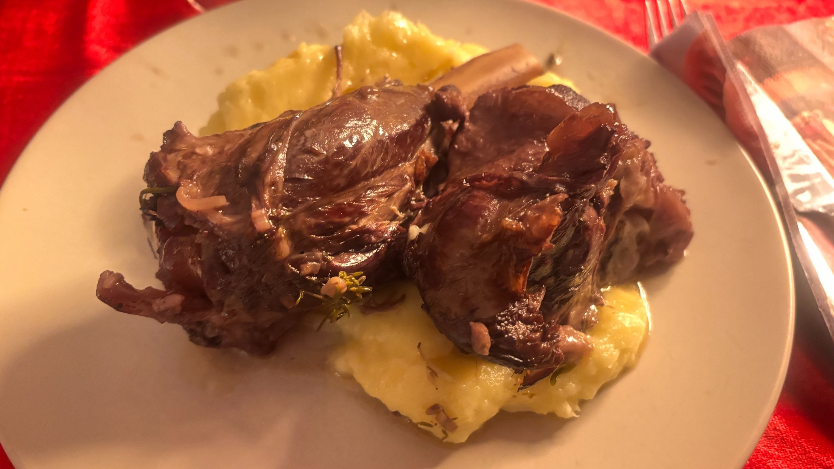 Braised lamb shank served on creamy mashed potatoes on a white plate, set on a red tablecloth.