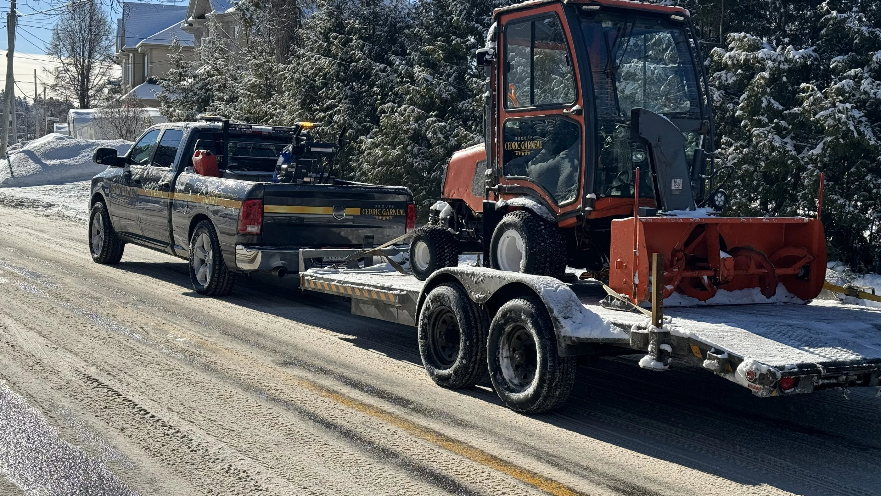 GCG pickup truck towing a trailer with a snow blower tractor on a snowy road.
