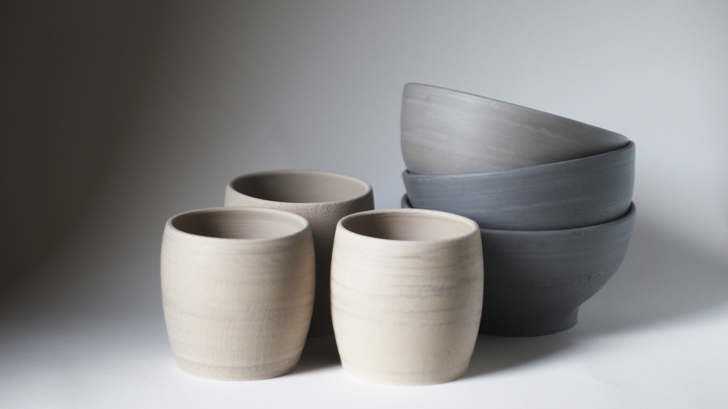 Trys smėlio spavos žiesti rankų darbo neglazūruoti puodeliai, šalia jų trys pilki bliūdukai