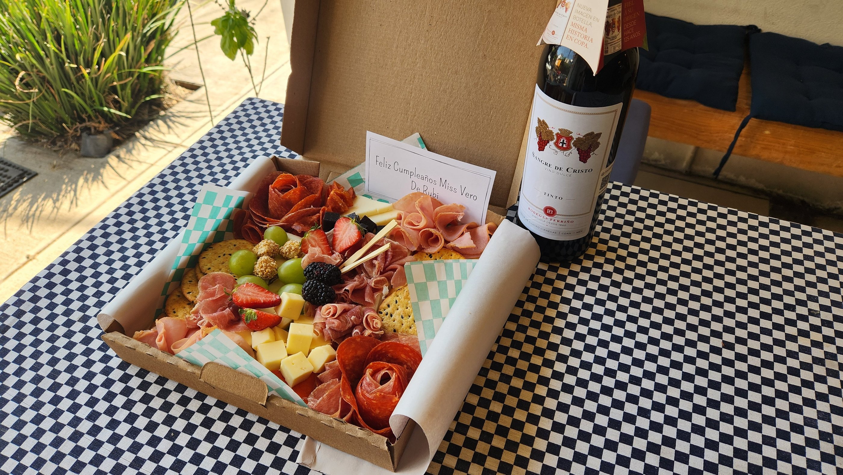 Gourmet charcuterie board gift box with cured meats, cheese, and fruit next to a bottle of red wine.