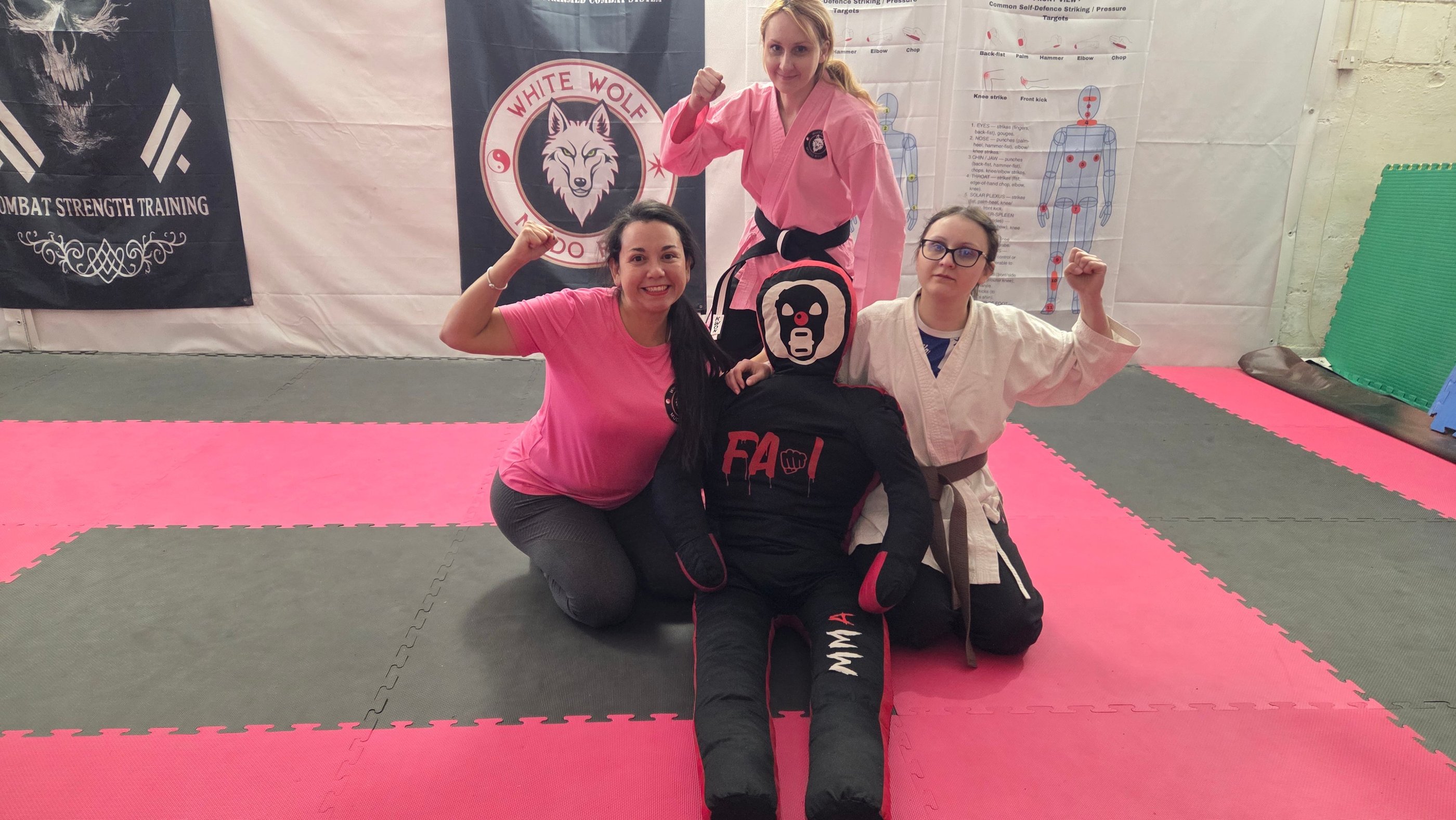Three female martial artists posing with an MMA training dummy on a pink gym mat.
