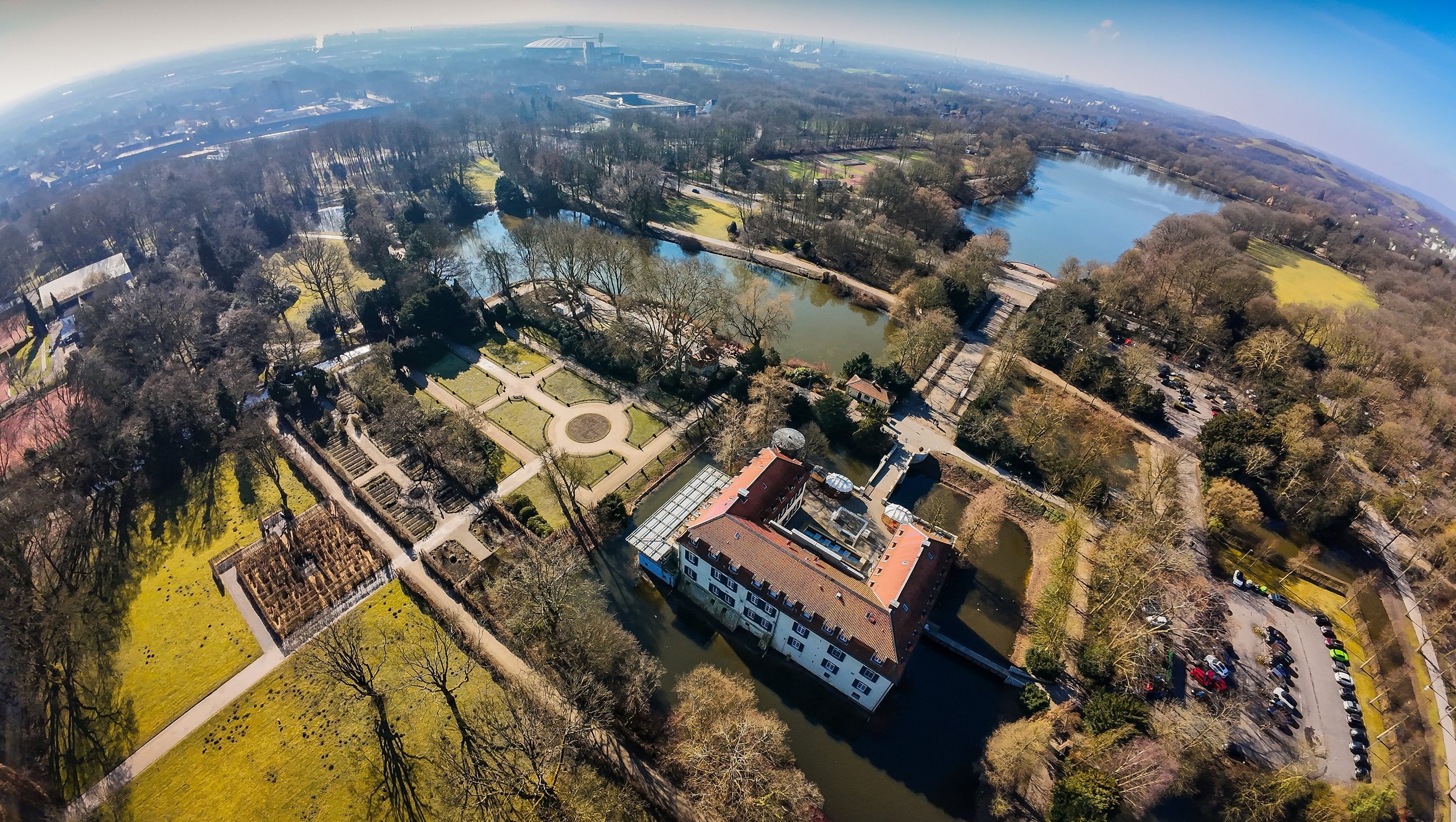 Aerial view of a historic moated castle surrounded by lush gardens, walking paths, and scenic lakes in a city park.