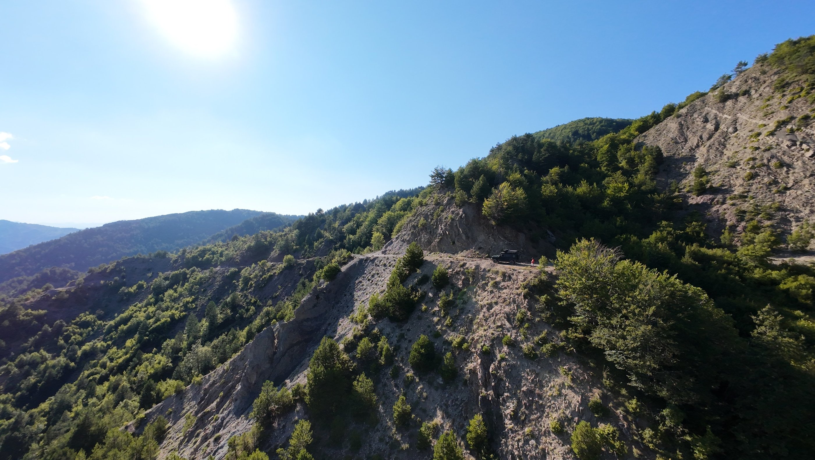 Off-road vehicle parked on a steep cliffside mountain trail overlooking a lush green forest valley.
