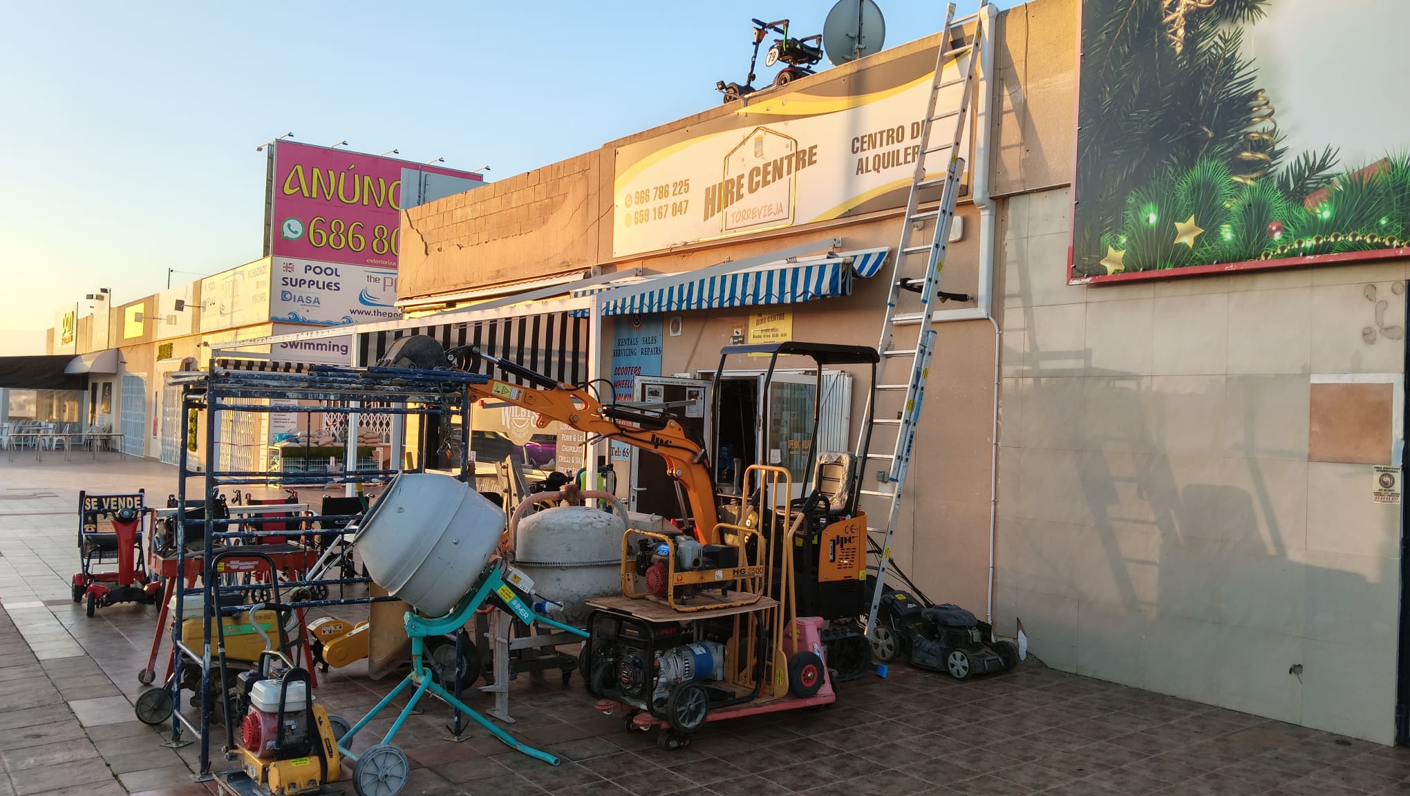Concrete mixer and mini excavator parked outside a construction equipment hire centre storefront.