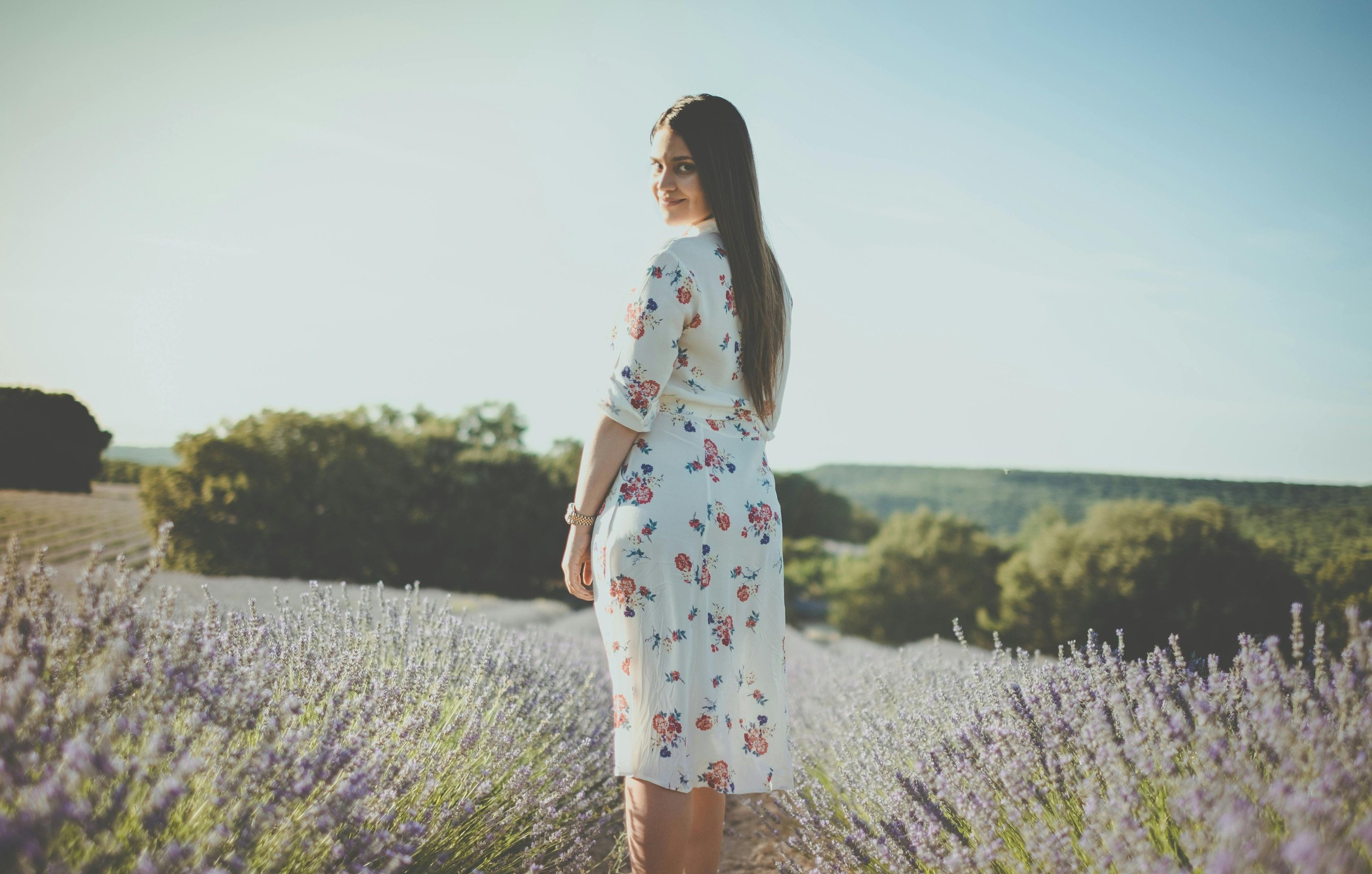 a woman in a white floral dress standing in a field of lavender. She looks at you and smiles.