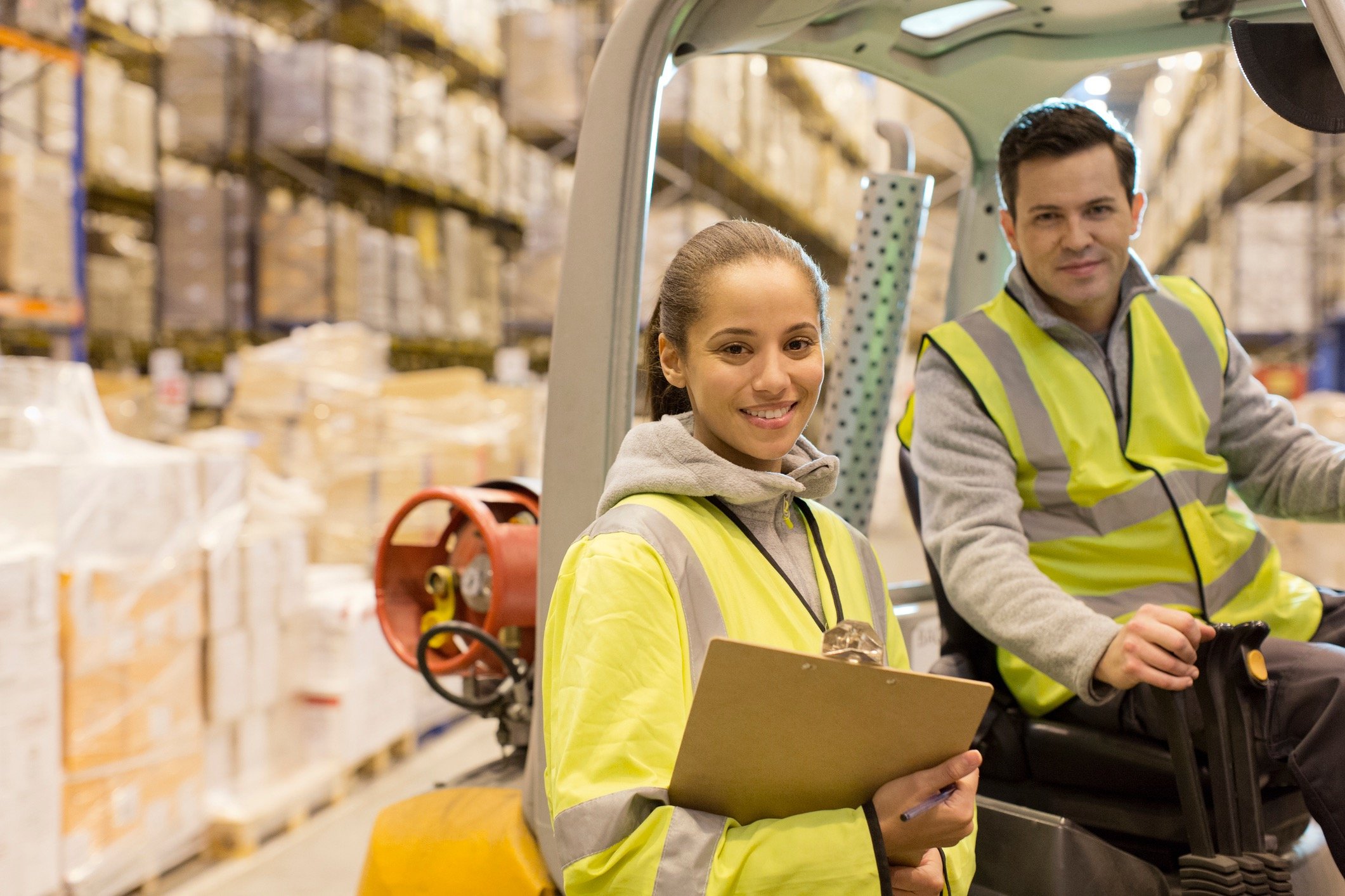 Trabajadores en un almacén usando chalecos reflectantes; uno manejando un montacargas y el otro con 