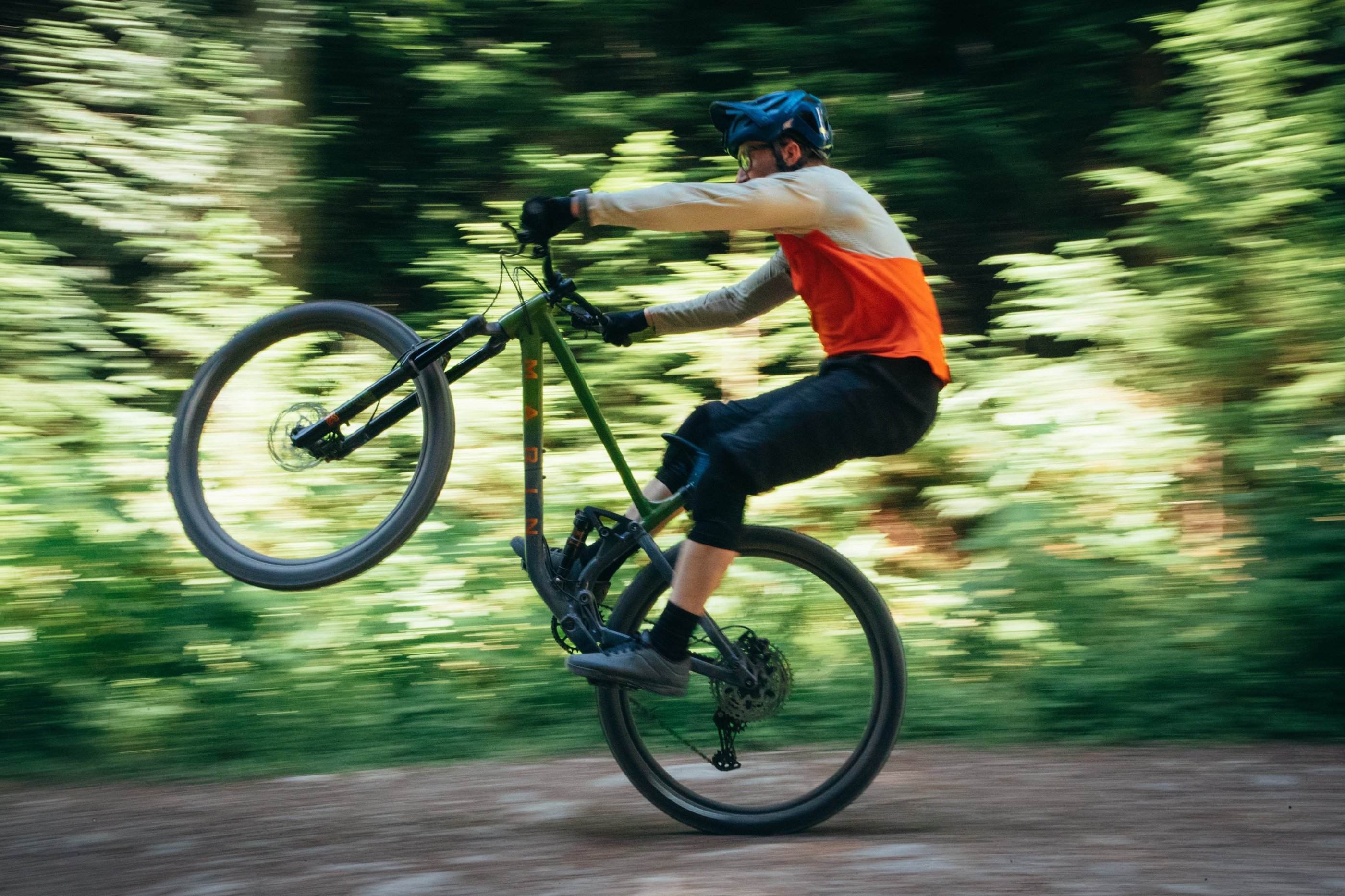 Mountain biker performing a manual wheelie on a green Marin bike through a sunlit forest trail.