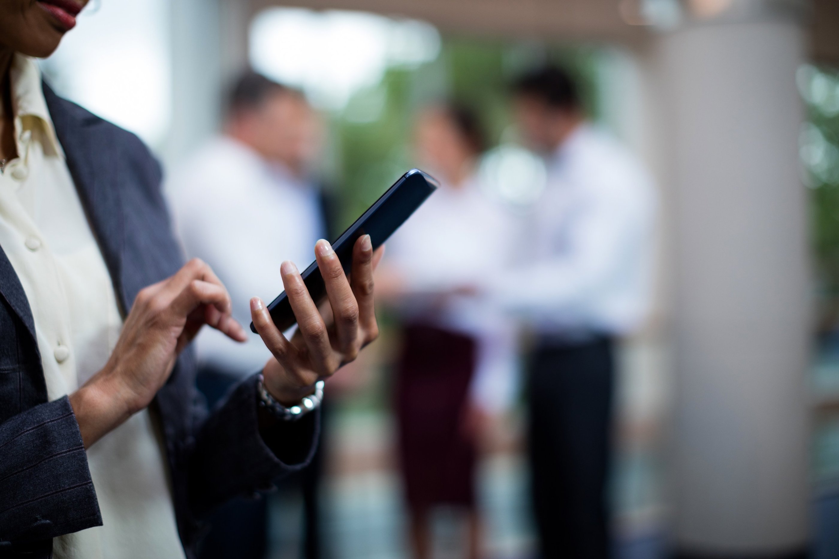 Person using a smartphone with colleagues conversing in the background.