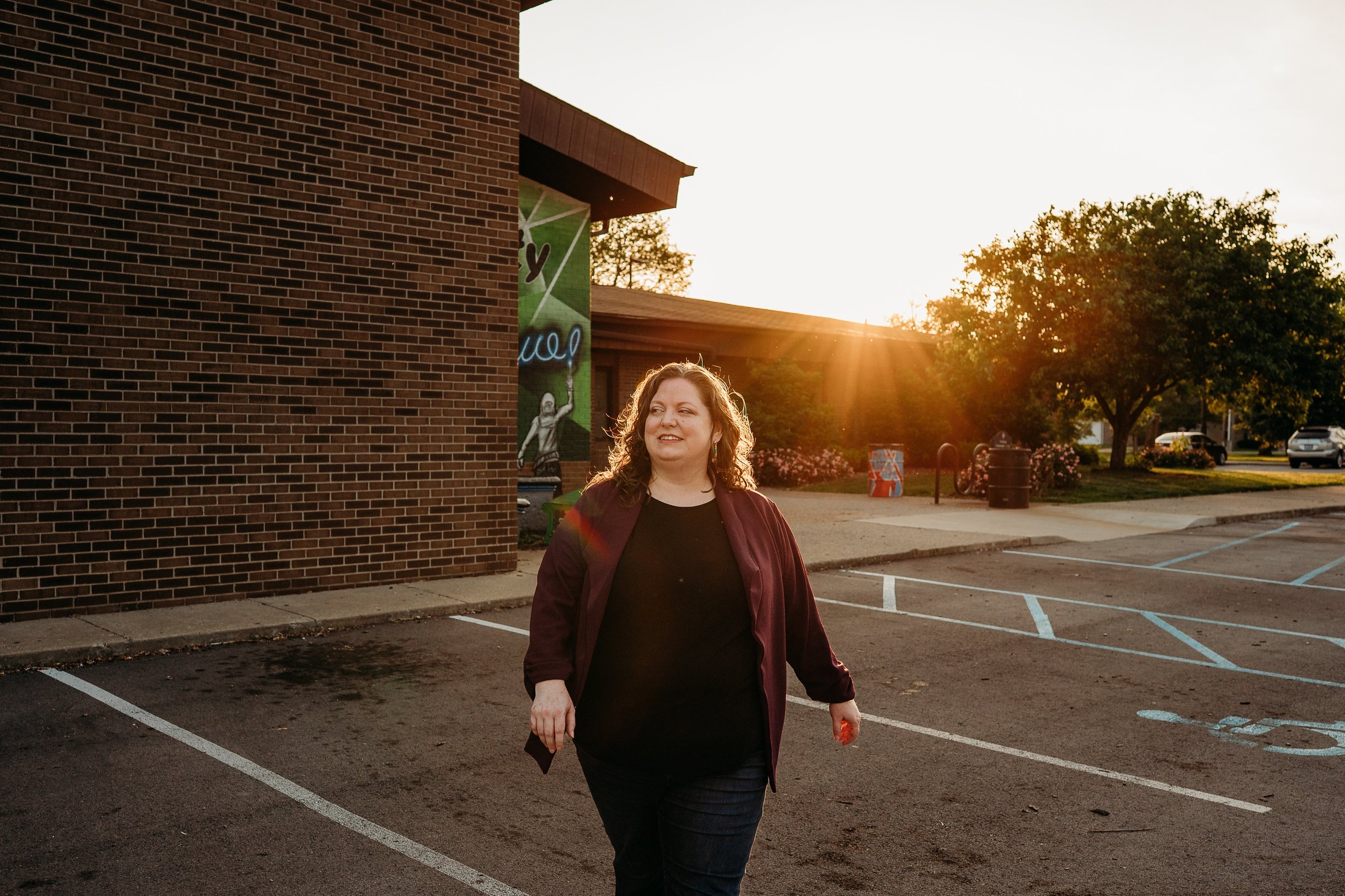 Smiling woman walking in a brick building parking lot at sunset with golden hour lighting.