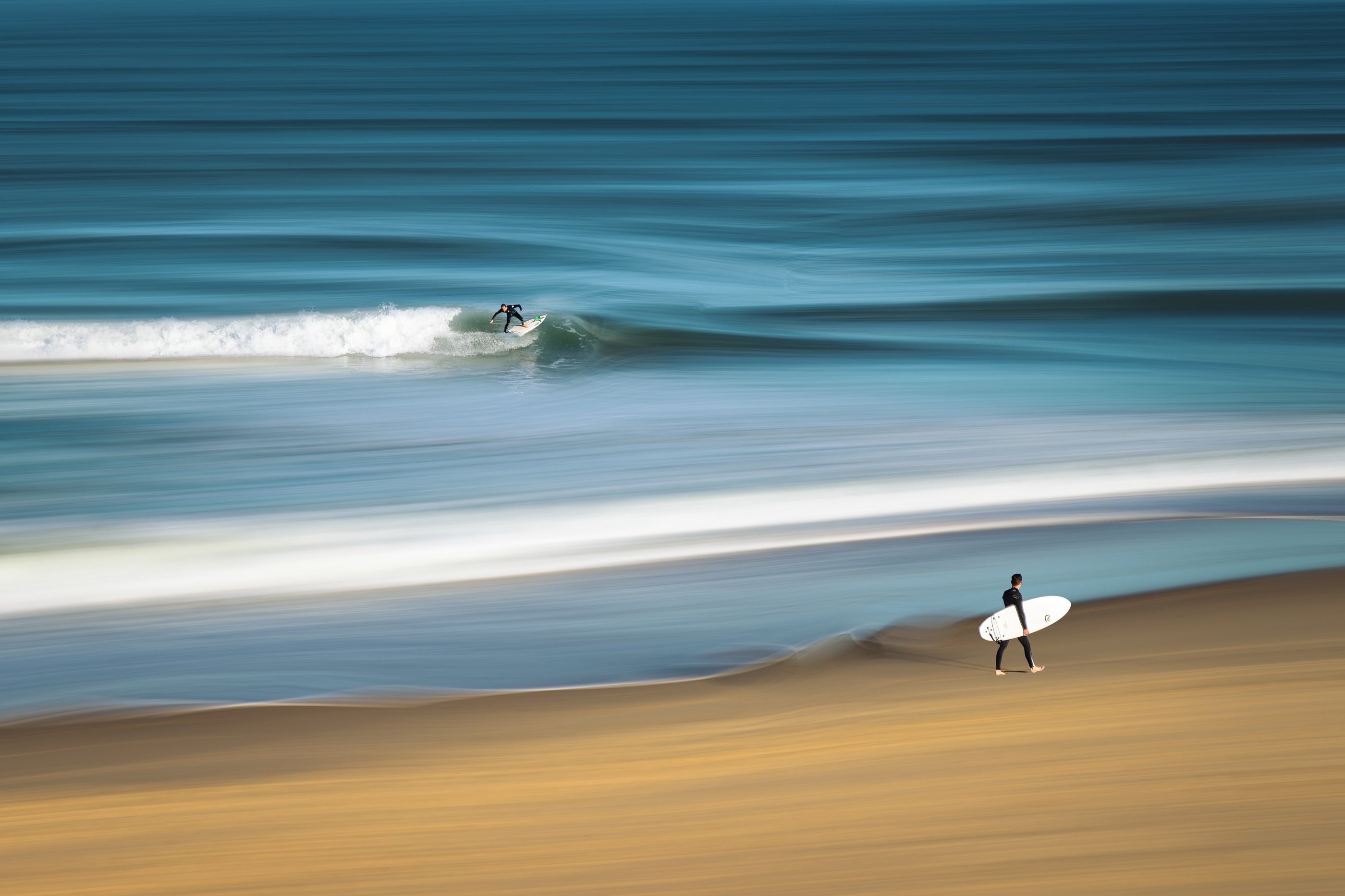 A long exposure motion blur of a surfer riding a blue ocean wave while another walks on the beach.