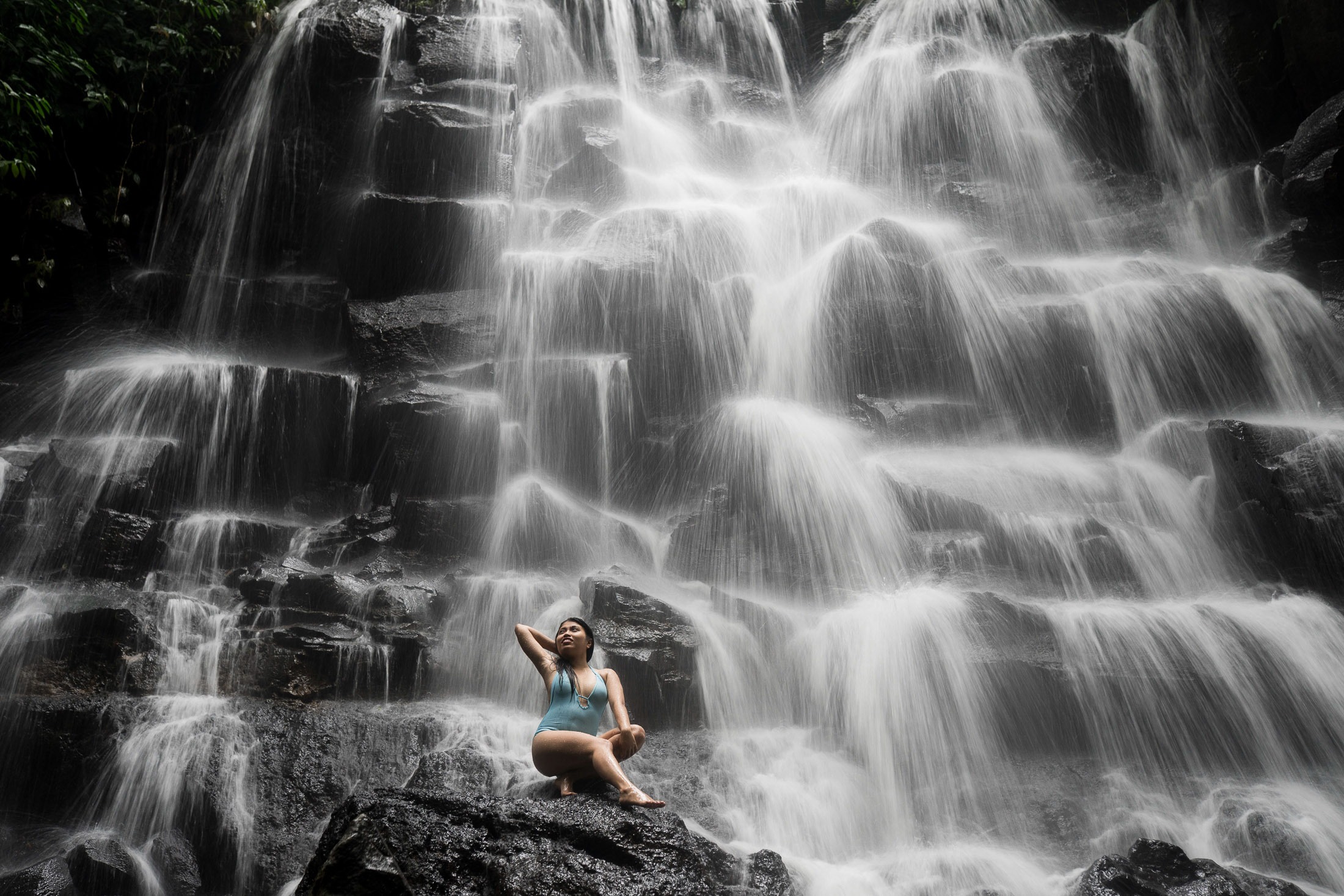 Wide portrait of woman sitting on rocks with waterfall at Kanto Lampo Bali