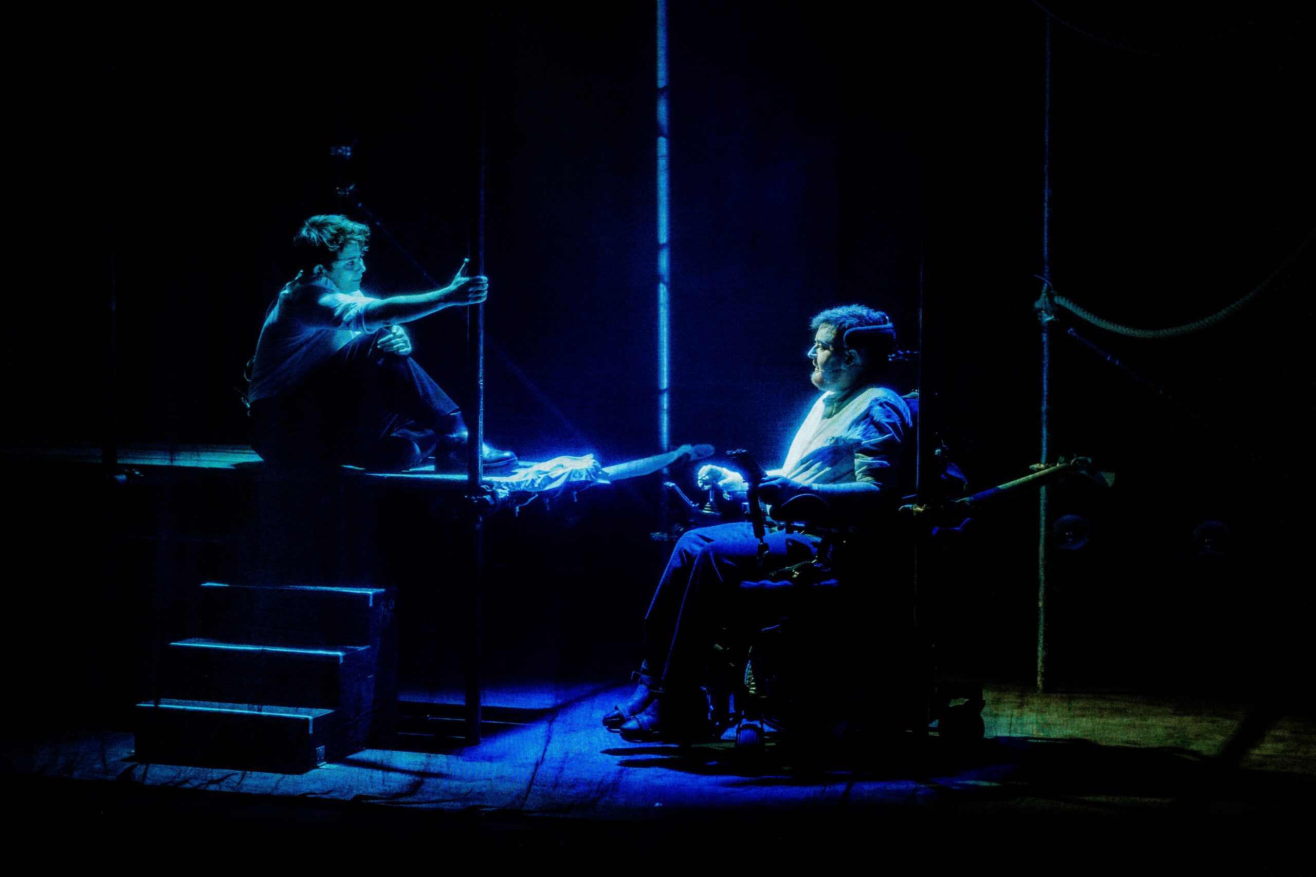 Soaked in blue light, Jamie Hale and CN Lester look at each other, surrounded by scaffolding