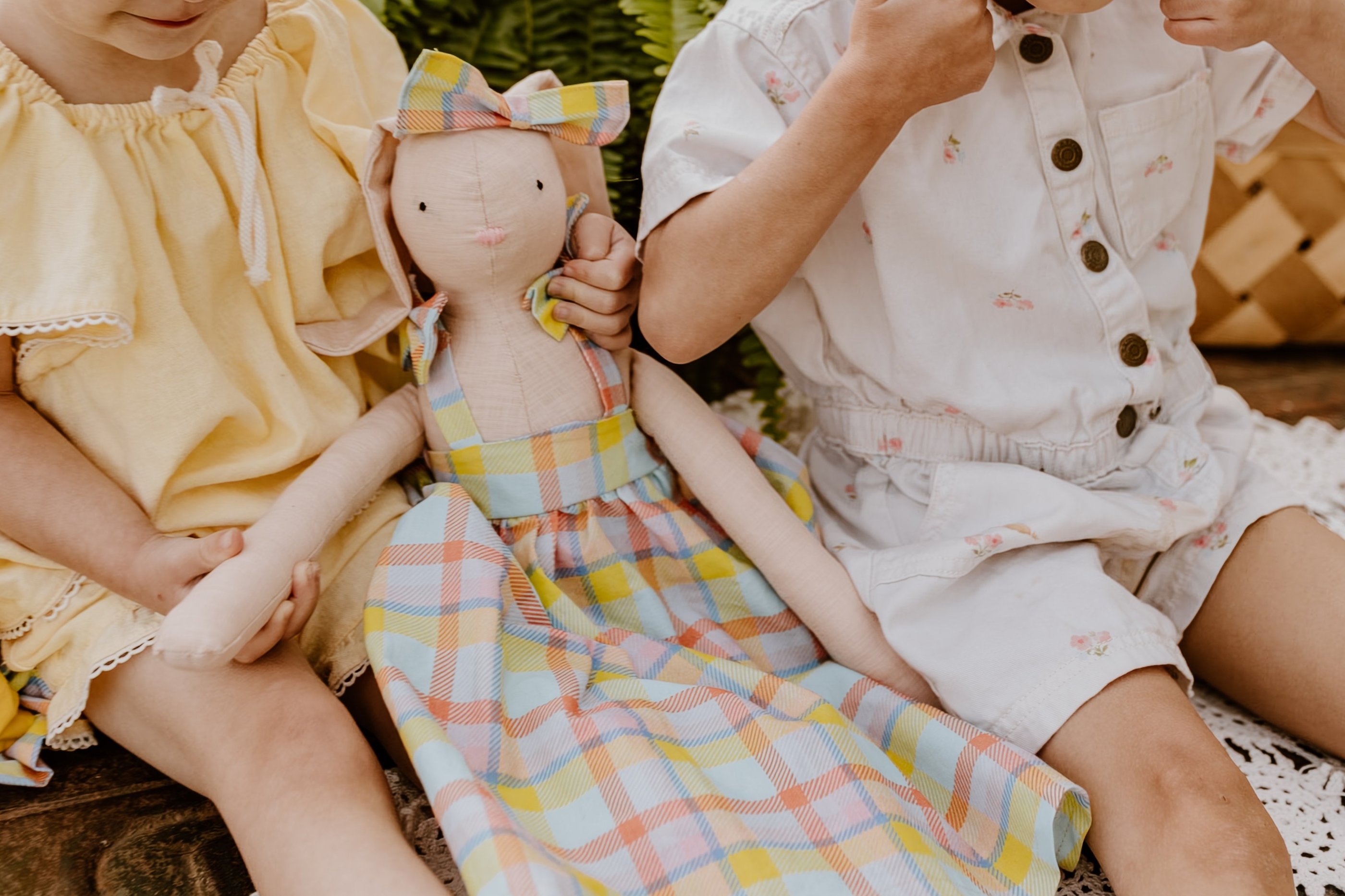 Two young children sitting together holding a handmade plush bunny doll wearing a colorful plaid dress.