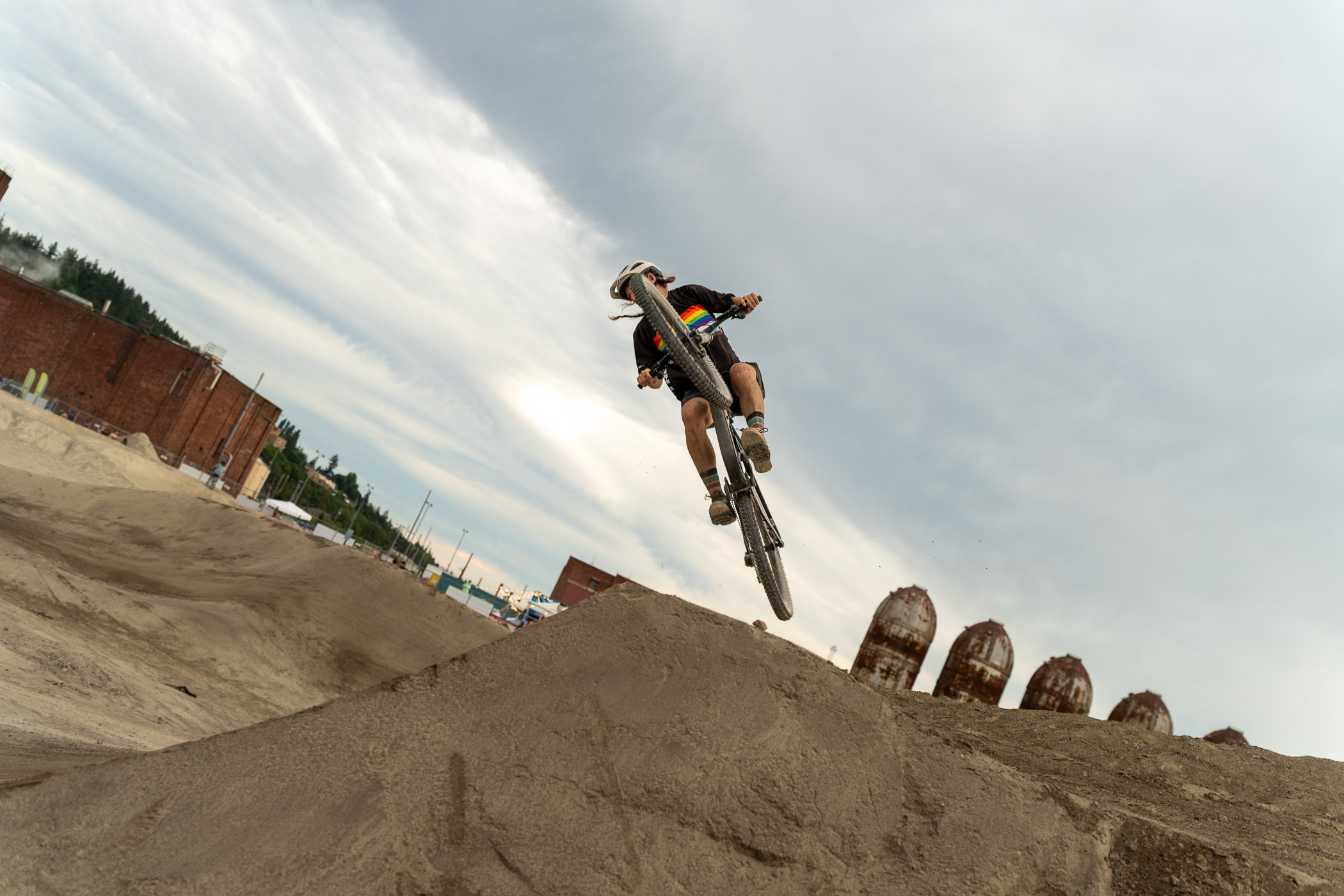 A mountain biker catches air off a dirt jump at an industrial bike park under a cloudy sky.