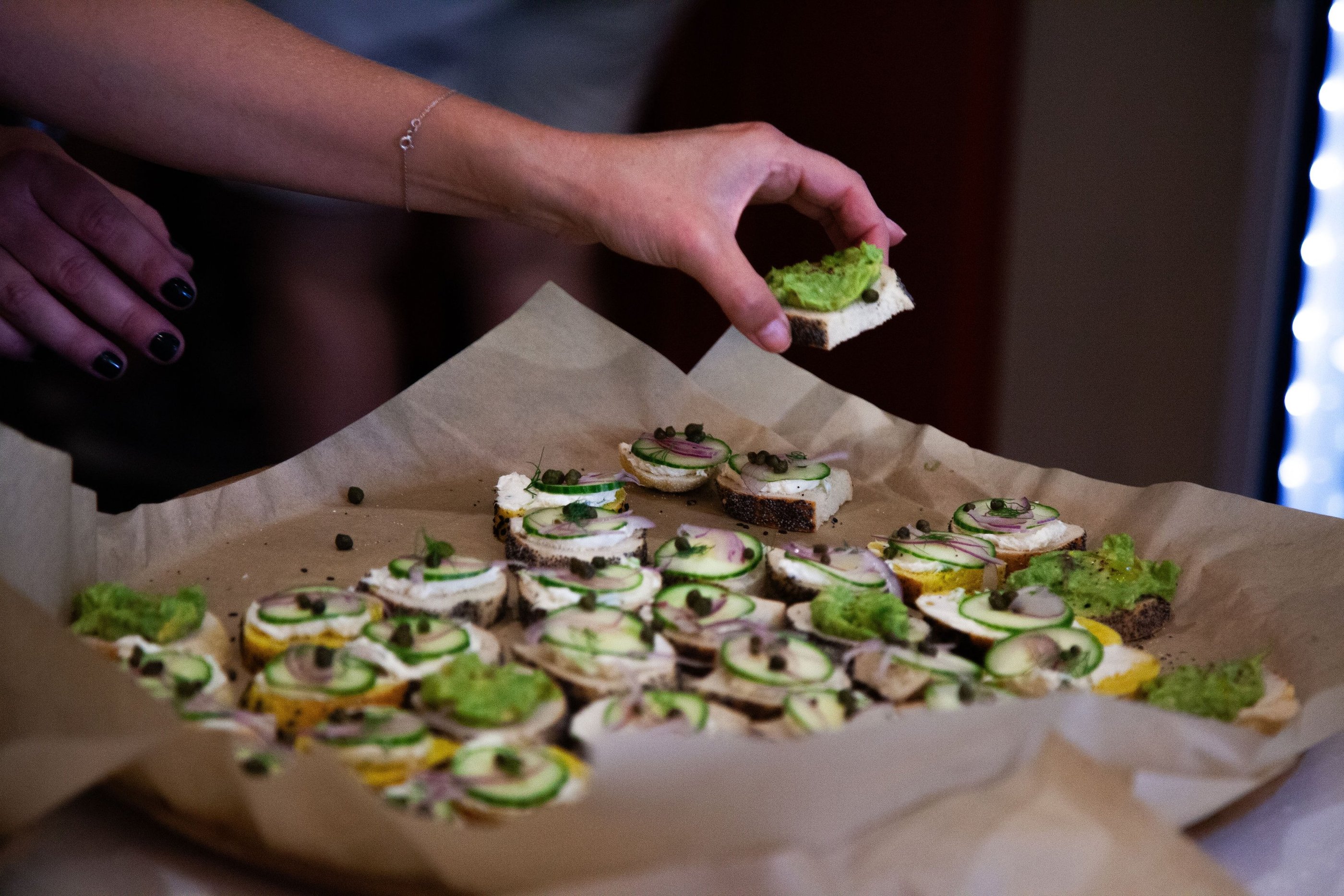 A hand picking up a small piece of bread with avocado on top from a large tray of similar snacks