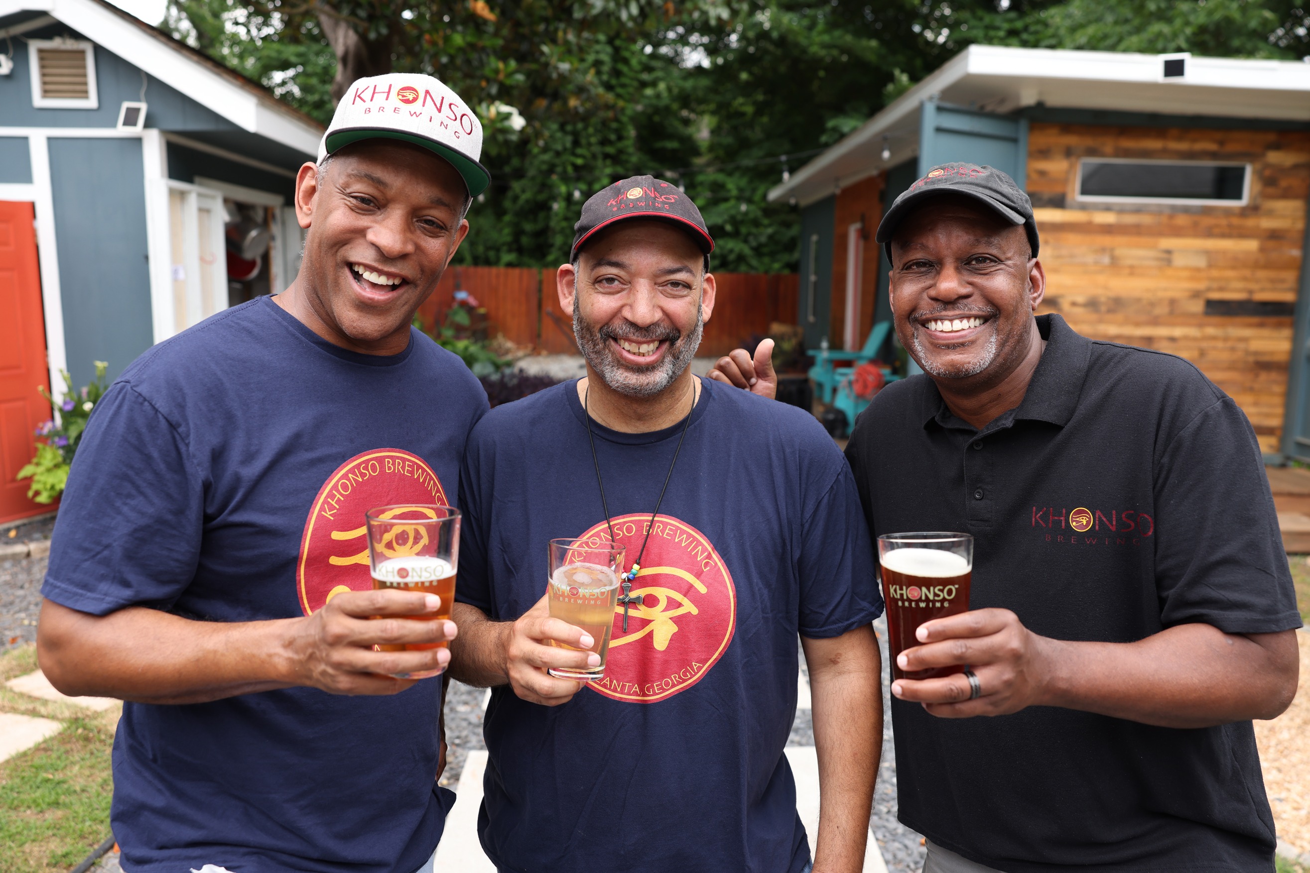 Three smiling men toast with glasses of craft beer while wearing Khonso Brewing apparel outdoors.